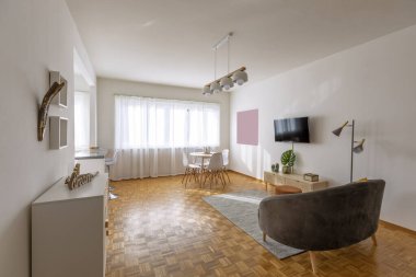 Oturma odasında halının üzerinde kumaş kanepe, duvarında televizyon asılı dolap ve beyaz sandalyeli masa. Ahşap zemin. İçeride kimse yok.