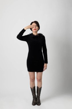 Beyaz arka planda seksi ve zarif siyah elbisesi olan güzel bir kadın sağ eliyle bir gözünü kapatıyor. Portre ve reklam resmi.