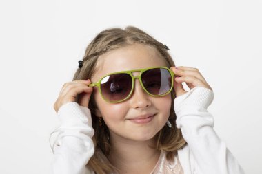 Little young teen girl, posing mischievously in the studio. The background is white and she has green sunglasses that are really too big for her.