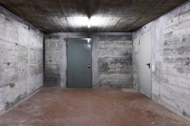 Zırhlı kapısı kapalı bir sığınağın güçlendirilmiş beton duvarının önü. Sahne beyaz bir neon lambayla aydınlatıldı. İçeride kimse yok.