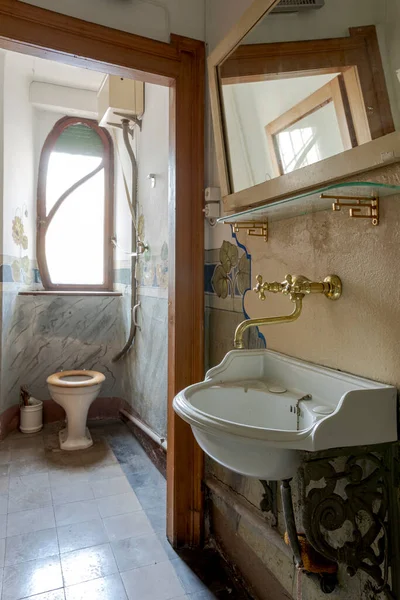 In a small room there is the toilet with the broken toilet seat, outside the door there is the sink with the faucet in gold. Interior of an abandoned villa to be restored