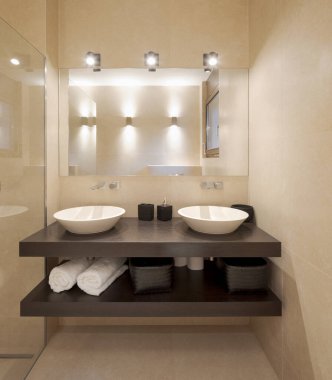 Geniş mermer levhalarla modern banyo manzarası. Sahne ışıklarıyla aydınlatılmış büyük aynalı iki lavabo var. İçeride kimse yok.