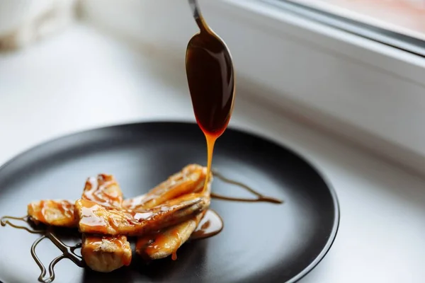 Čajová lžička kape karamelizovaný sirup na banán na černý talíř, bílý stůl, veganský dezert. Selektivní zaměření Stock Fotografie