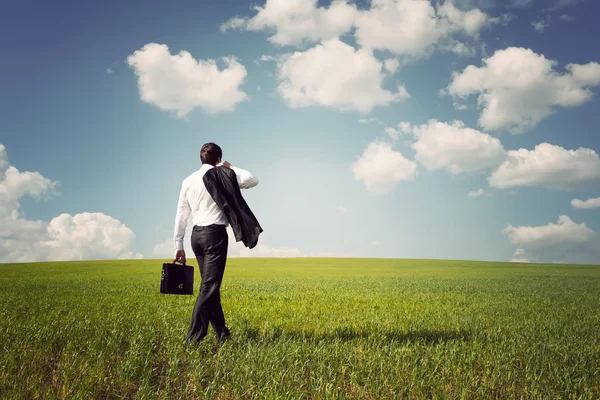 Businessman in a suit walking on a spacious green field with a b — Stock Photo, Image