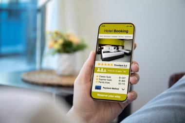 Erkek eller telefonu tutuyor ve ekranda otel rezervasyonu var. 