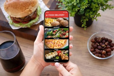 Kadın eli ahşap masanın üzerinde yemek dağıtım uygulaması olan telefonu tutuyor. 