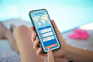 Kadın elleri başvuru ile telefonu tutar. Yaz aylarında arka plandaki havuzda uçak biletlerini ararlar.