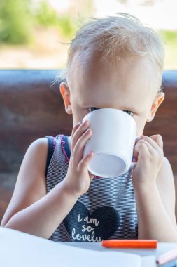 Elinde bir bardak süt olan bir bebek ahşap bir evin terasında oturuyor.