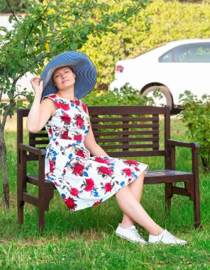 Çiçekli elbise ve şapka giymiş bir kadın bahçede bir bankta oturuyor.
