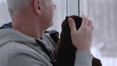 Yaşlı bir adam ve kedisi pencereden dışarı bakıyor.