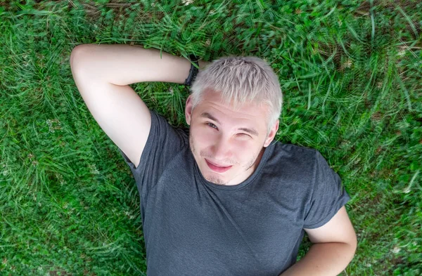 Ein fröhlicher Typ im grauen T-Shirt liegt mit zusammengekniffenen Augen auf einem grünen Rasen. — Stockfoto