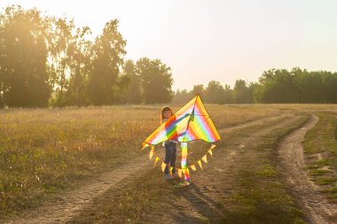 Yaz tarlasında kır yolunda koşan gökkuşağı renkli uçurtmalı küçük bir kız. Seçici odak.