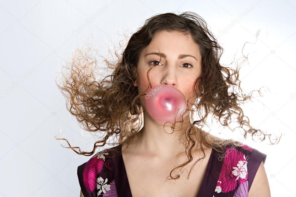 Close up portrait of a young woman blowing a strawberry bubble gum while her hair flies in the air, isolated against a plain background.