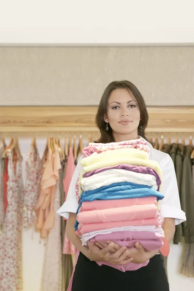 Asistente de tienda sosteniendo una pila de ropa en una tienda de moda . — Foto de Stock