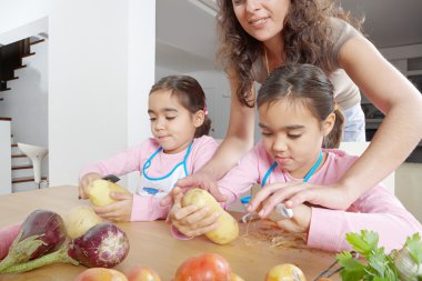Mother teaching twin daughters to peel potatoes in the kitchen using a chopping board and peelers. clipart