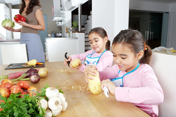 Mutter und Zwillingstöchter lernen gemeinsam in der Küche Kartoffeln schälen, mit einem Schneidebrett mit Obst und Gemüse. — Stockfoto