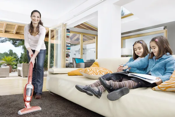 Joven madre usando un limpiador de vacum con sus dos hijas gemelas mirar un libro en la sala de estar . — Foto de Stock