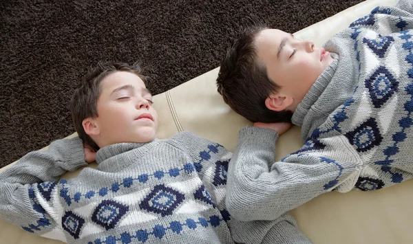 Dos hermanos gemelos idénticos durmiendo en un sofá de cuero blanco con jerséis idénticos . —  Fotos de Stock
