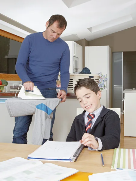 Vater und Sohn machen Hausaufgaben und bügeln Kleidung, während sie zu Hause in der Küche stehen. — Stockfoto