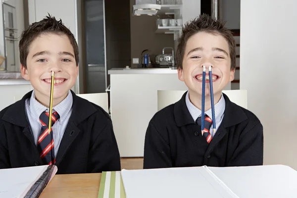 Dos hermanos gemelos idénticos jugando juegos divertidos mientras hacen su tarea en casa en la mesa de la cocina . — Foto de Stock