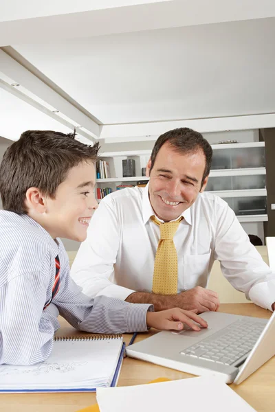Vater hilft seinem Sohn bei den Hausaufgaben, während er sich zu Hause einen Laptop teilt und lächelt. — Stockfoto