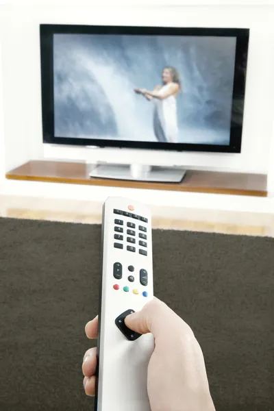 Mano dell'uomo che tiene un telecomando della TV, premendo un pulsante mentre punta ad una TV a schermo piatto. — Foto Stock