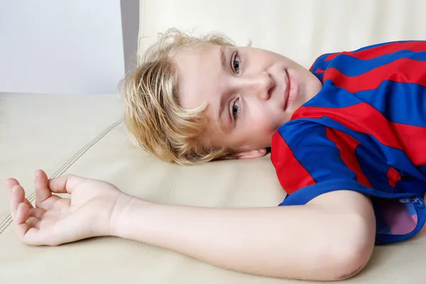 Niño acostado en un sofá de cuero blanco sonriendo y vistiendo una camisa de fútbol de rayas rojas y azules . — Foto de Stock