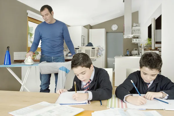 Two twin brothers doing homework on kitchen table while dad irons clothes. — Stock Photo, Image