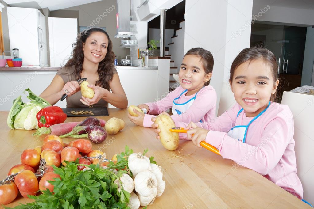 Mother and twin daughters learning to peel potatoes together in the kitchen, using a chopping board with fruit and vegetables.