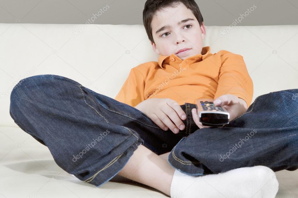 Young boy using a tv remote control while watching television sitting on a white leather sofa at home.