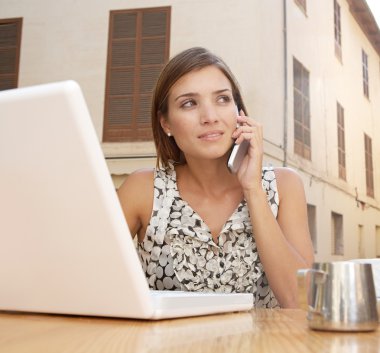 açık havada bir kafede masada oturarak ise bir cep telefonu ve dizüstü bilgisayar kullanarak iş kadını.