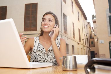 açık havada bir kafede masada oturarak ise bir cep telefonu ve dizüstü bilgisayar kullanarak bir işkadını portresi.