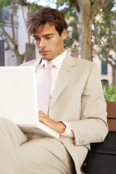 Attraente uomo d'affari seduto su una panchina di legno in una città classica utilizzando un computer portatile . — Foto Stock
