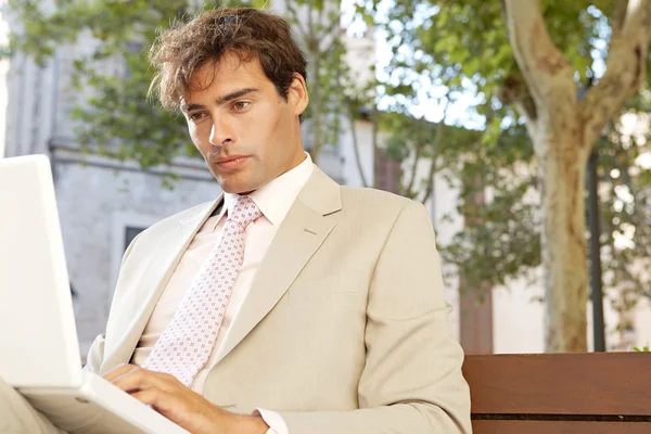 Attraktive Geschäftsmann sitzt auf einer Holzbank in einer klassischen Stadt mit einem Laptop-Computer. — Stockfoto