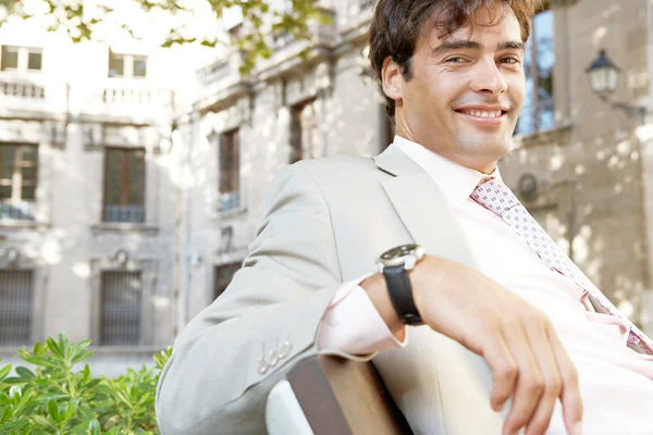 Porträtt av en ung attraktiv affärsman som sitter på en träbänk i en klassisk stad, leende. — Stockfoto