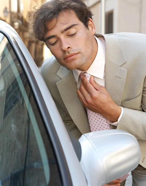 Ελκυστική νεαρός επιχειρηματίας περιποίηση χρησιμοποιώντας ένα αυτοκίνητο οπισθοπορείας καθρέφτη να βάλει τάξη του γραβάτα κόμπος — Φωτογραφία Αρχείου