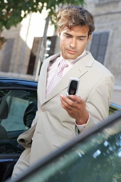 Empresário usando um telefone celular para enviar uma mensagem enquanto está em pé por alguns carros na cidade . — Fotografia de Stock