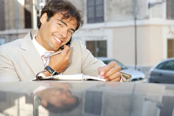 Nahaufnahme eines attraktiven Geschäftsmannes, der ein Handy benutzt und Notizen macht, während er sich in der Stadt an ein Auto lehnt — Stockfoto