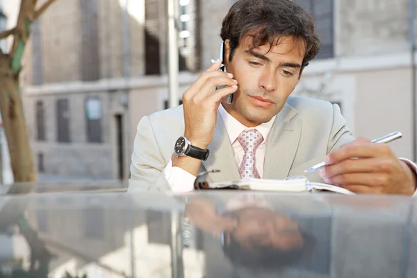 Attractive businessman using a cell phone and taking notes while leaning on a car in the city. — Stock Photo, Image