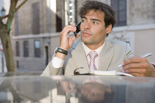 Homem de negócios profissional atraente usando um telefone celular e tomar notas enquanto em uma conversa chamada telefônica em um parque de estacionamento — Fotografia de Stock