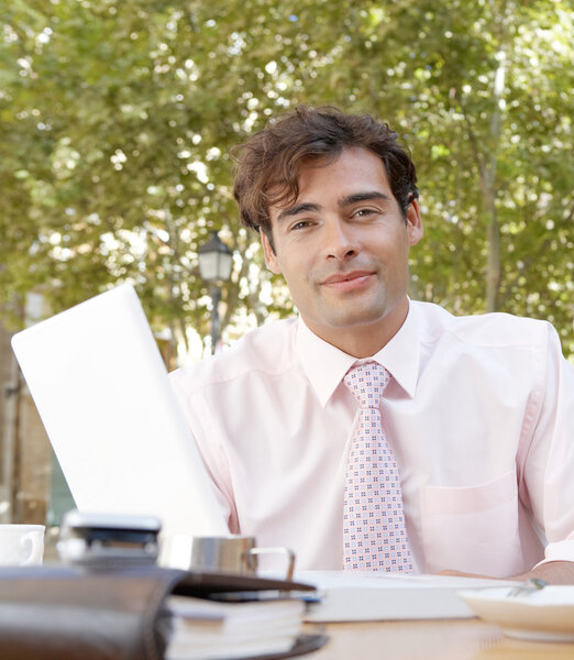 Businessman sitting at a coffee shop table using a laptop computer and smiling at camera.