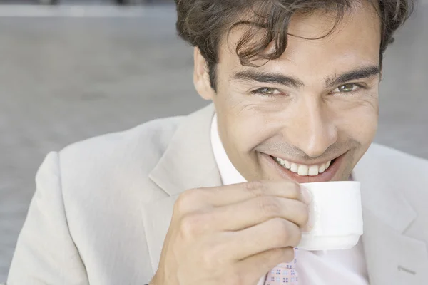 Atractivo joven empresario sonriendo a la cámara mientras bebe una taza de café al aire libre . — Foto de Stock