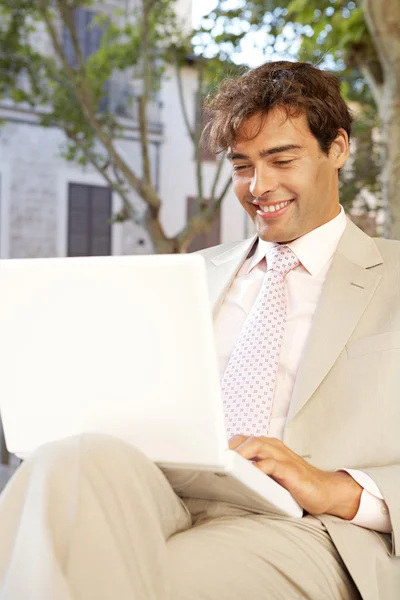 Attraktive Geschäftsmann sitzt auf einer Holzbank in einer klassischen Stadt mit einem Laptop-Computer und lächelt. Stockbild