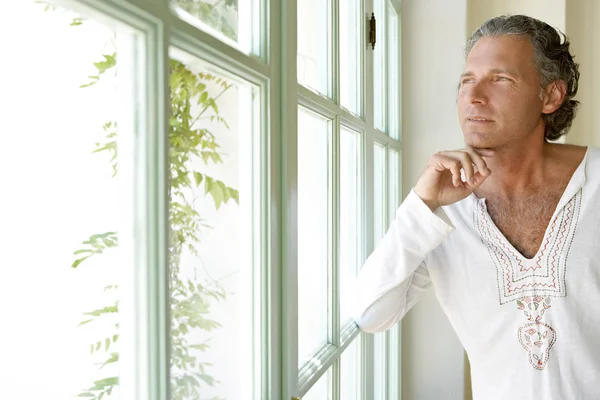 Зрелый мужчина смотрит из большого окна на дом, будучи вдумчивым . — стоковое фото