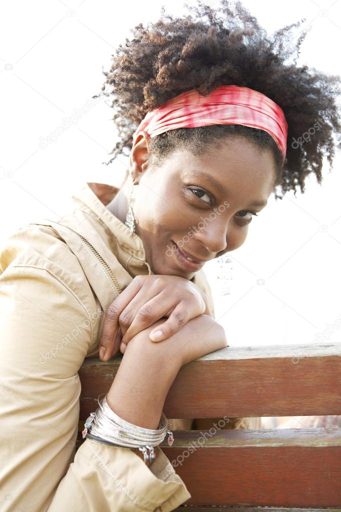 Young attractive african american woman smiling while sitting on a bench in a city park.