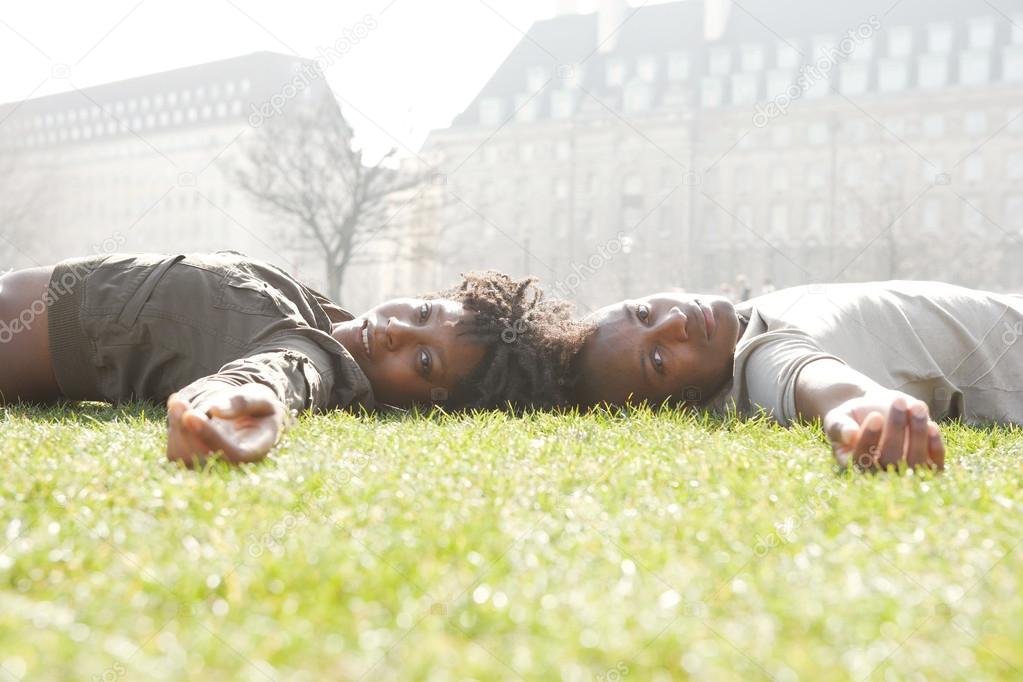Attractive black couple laying down on green grass head to head, while visiting London city on vacations.