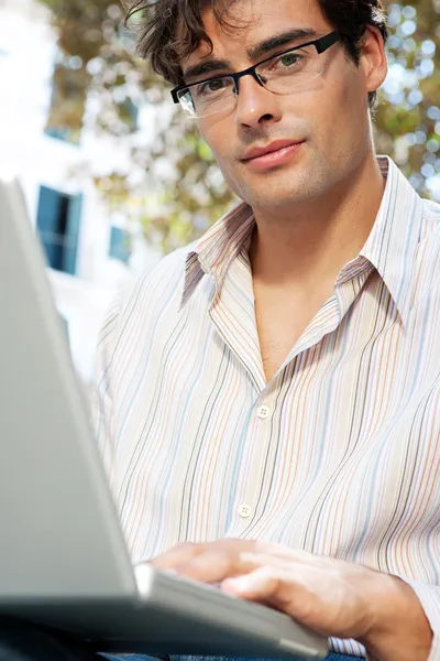 Joven hombre de negocios usando una computadora portátil mientras está sentado en un banco en un parque de la ciudad — Foto de Stock