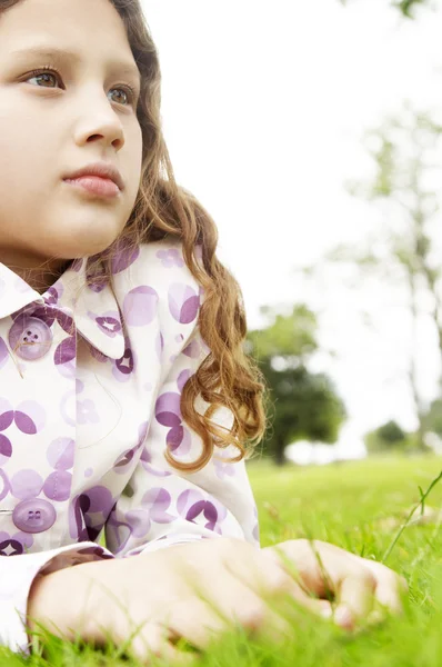 Portrait of a young girl laying down on green grass in the park, looking ahead. — Stock Photo, Image