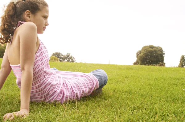 Side view of a girl sitting down on green grass in the park, being thoughtful. — Stock Photo, Image