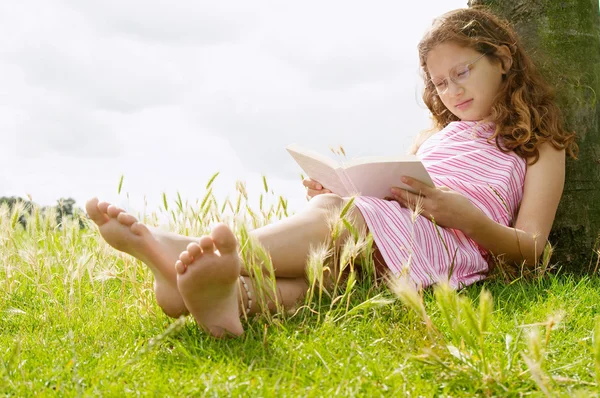 Ung flicka som läser en bok medan du sitter under ett träd i parken. — Stockfoto